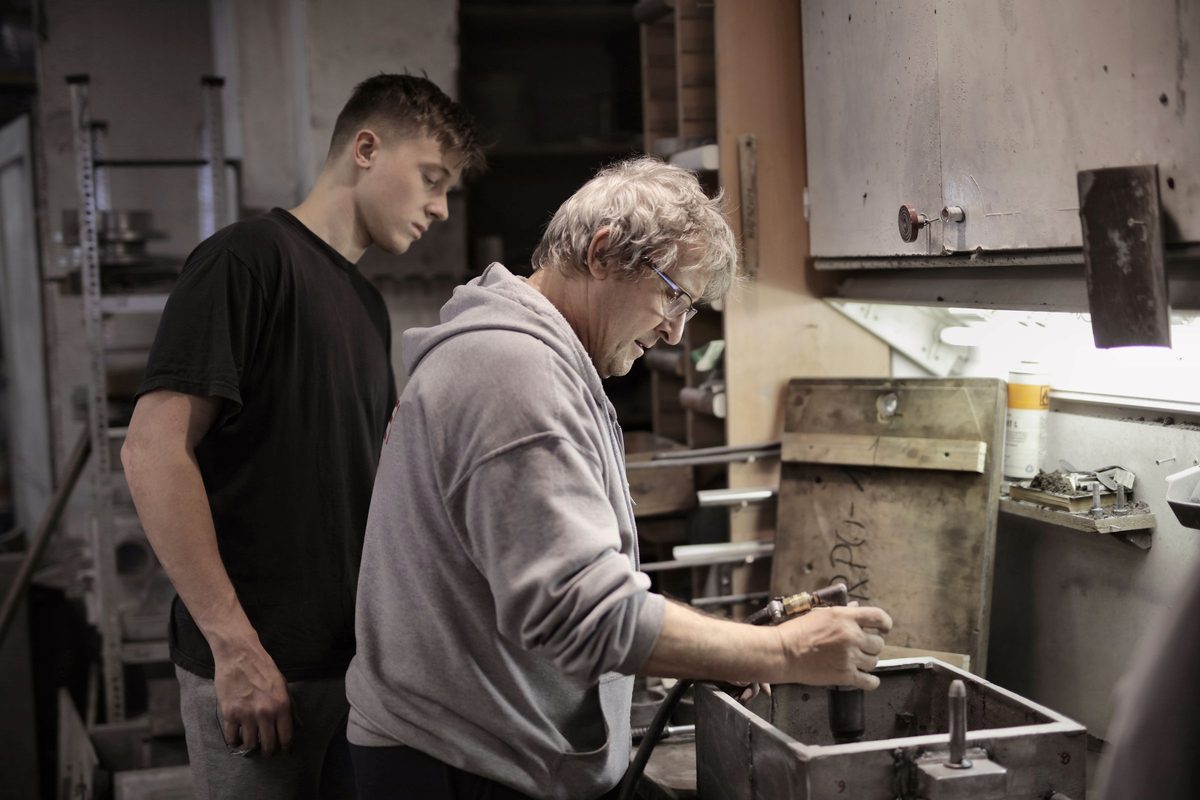 An experienced craftsman and a younger colleague working side by side at a bench in a workshop
