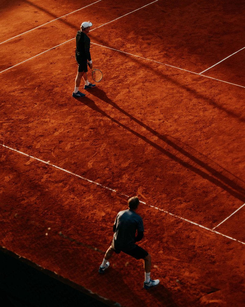 Two tennis players seen from above on a red clay court at sunset, one standing near the baseline holding a racket with a long shadow stretching behind him, the other crouched ready in the foreground