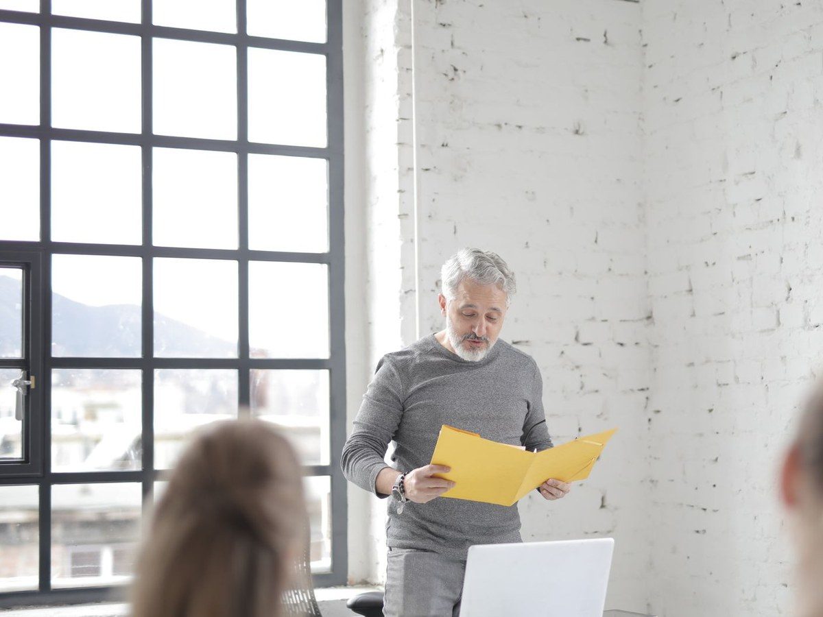 A silver-haired, bearded older man in a grey sweater holding a yellow folder and speaking with colleagues in a bright brick-walled workspace with large windows